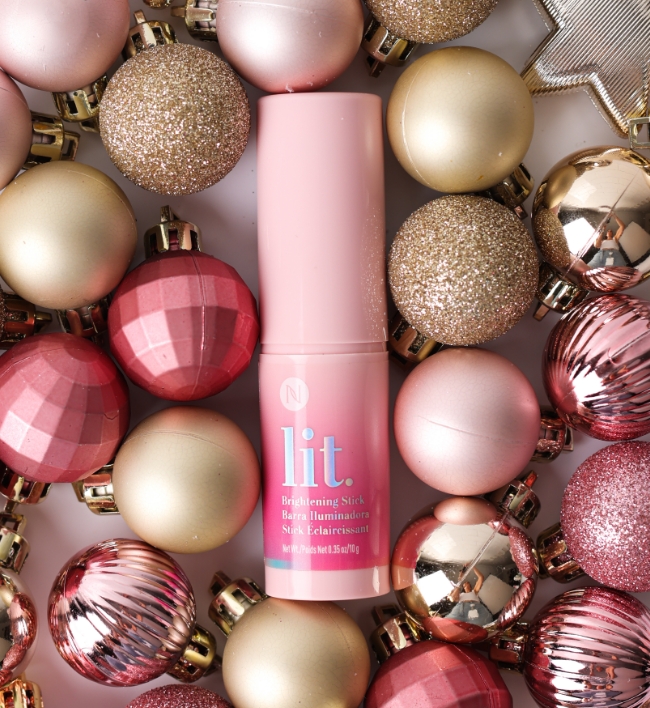 Lit Brightening Stick surrounded by pink and gold holiday ornaments. 